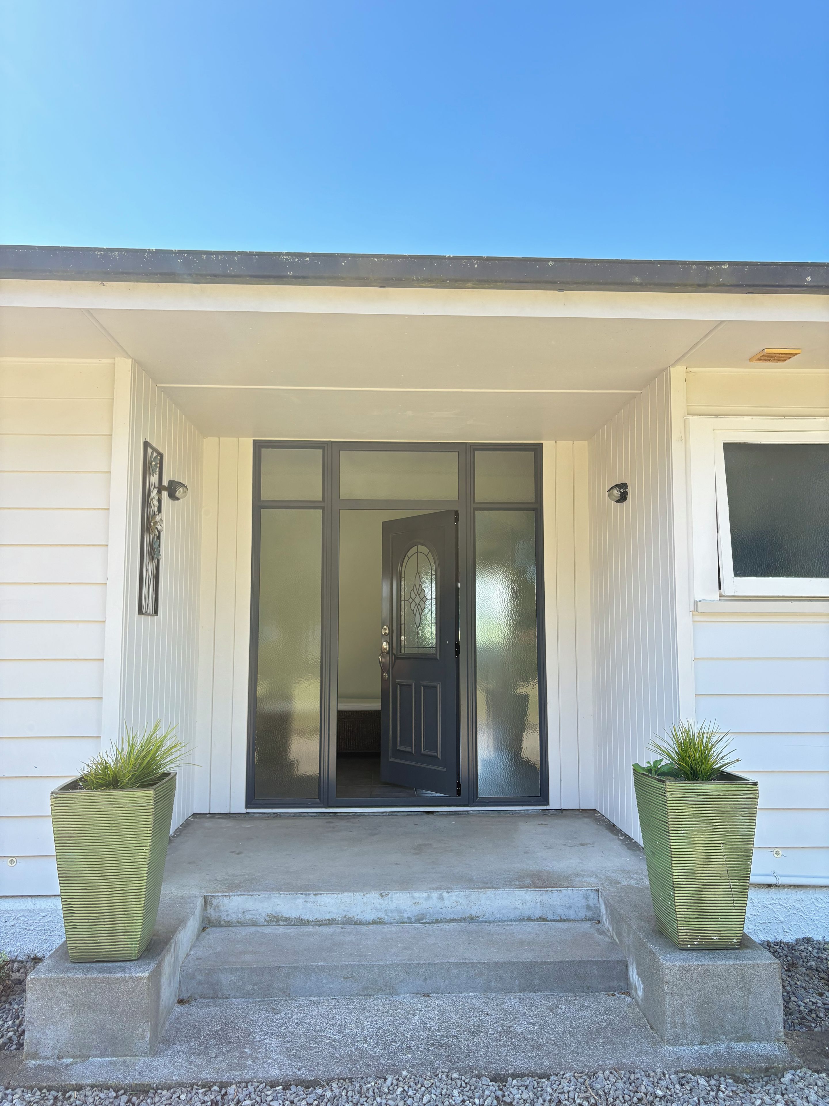 A house entrance with a dark door, white siding, and two green planters on the steps.
