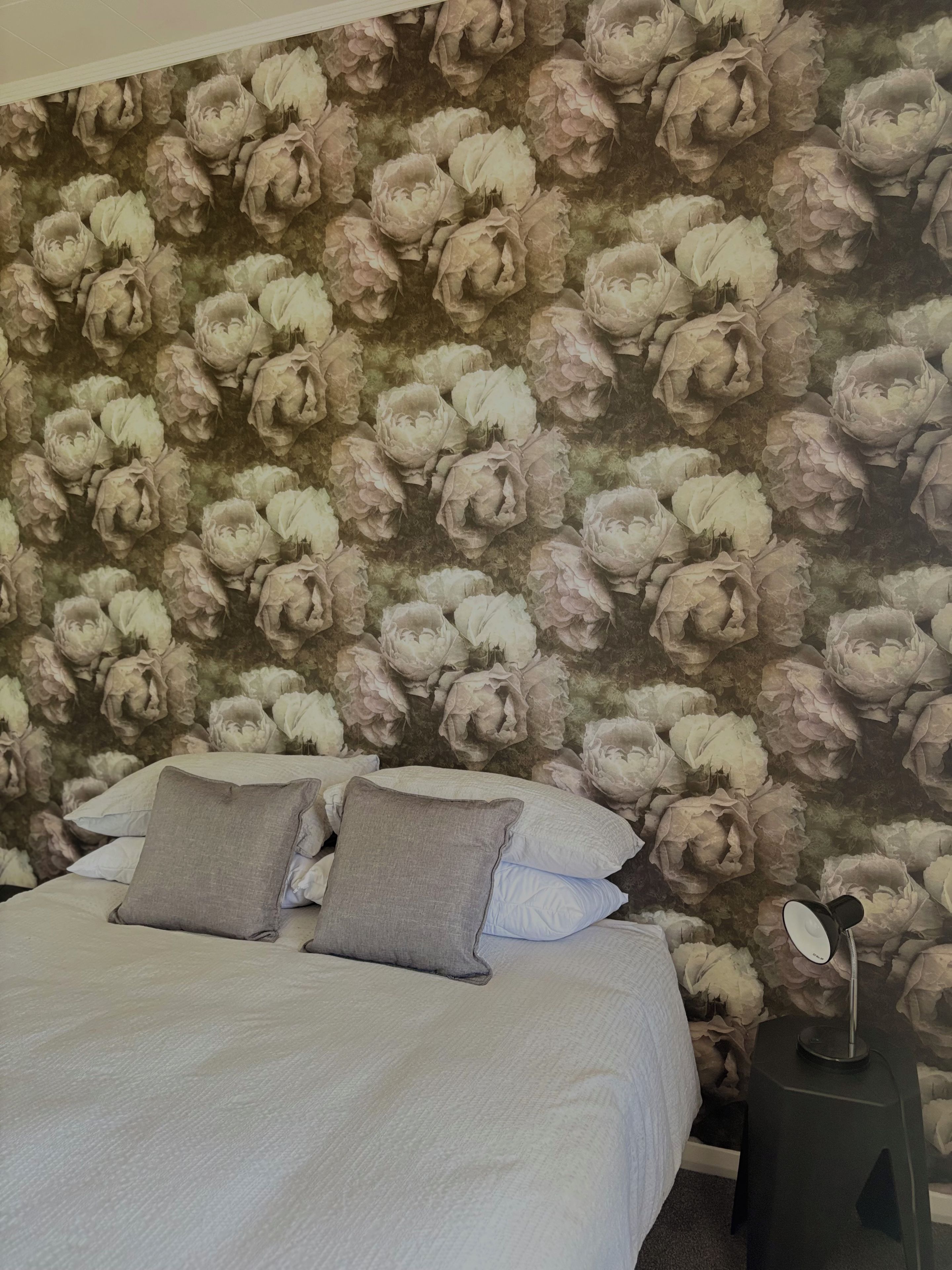 Quarto com papel de parede floral, cama com roupa de cama branca e duas almofadas, e mesa de cabeceira com abajur.