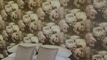Quarto com papel de parede floral, cama com roupa de cama branca e duas almofadas, e mesa de cabeceira com abajur.