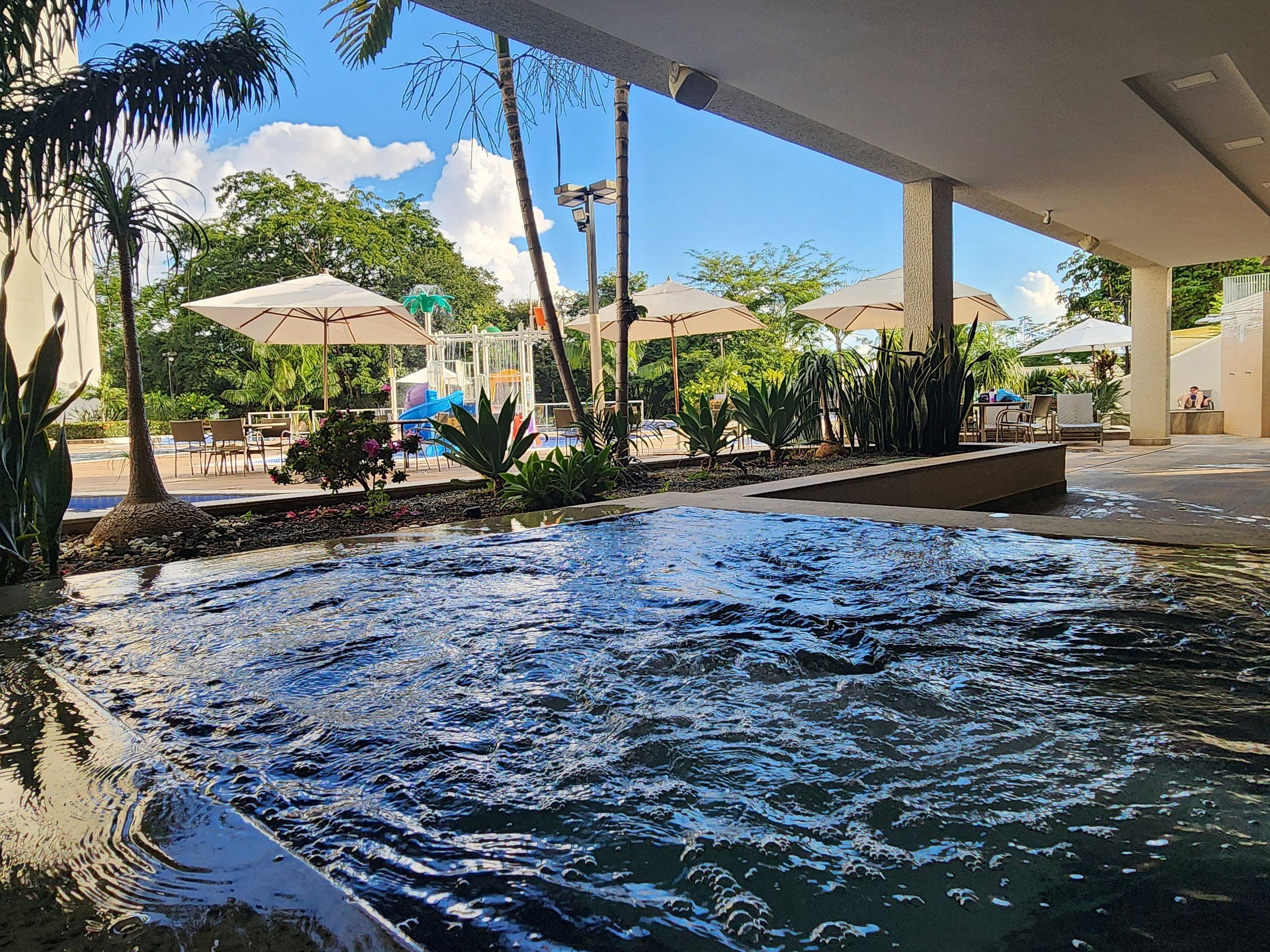 Área interna da piscina com um recurso aquático, cercada por palmeiras e guarda-sóis.