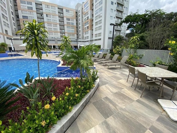 A swimming pool with lounge chairs and tables, surrounded by greenery and a building in the background. - Apto 2 Quartos p/ 8 - acesso ao rio (Rio Quente)