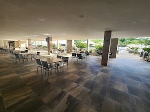 A spacious indoor area with multiple tables and chairs, a tiled floor, and a view of greenery outside. - Apto 2 Quartos p/ 8 - acesso ao rio (Rio Quente)
