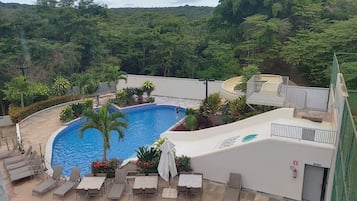 Una piscina con sillones de descanso y mesas, rodeada de vegetación y un edificio.