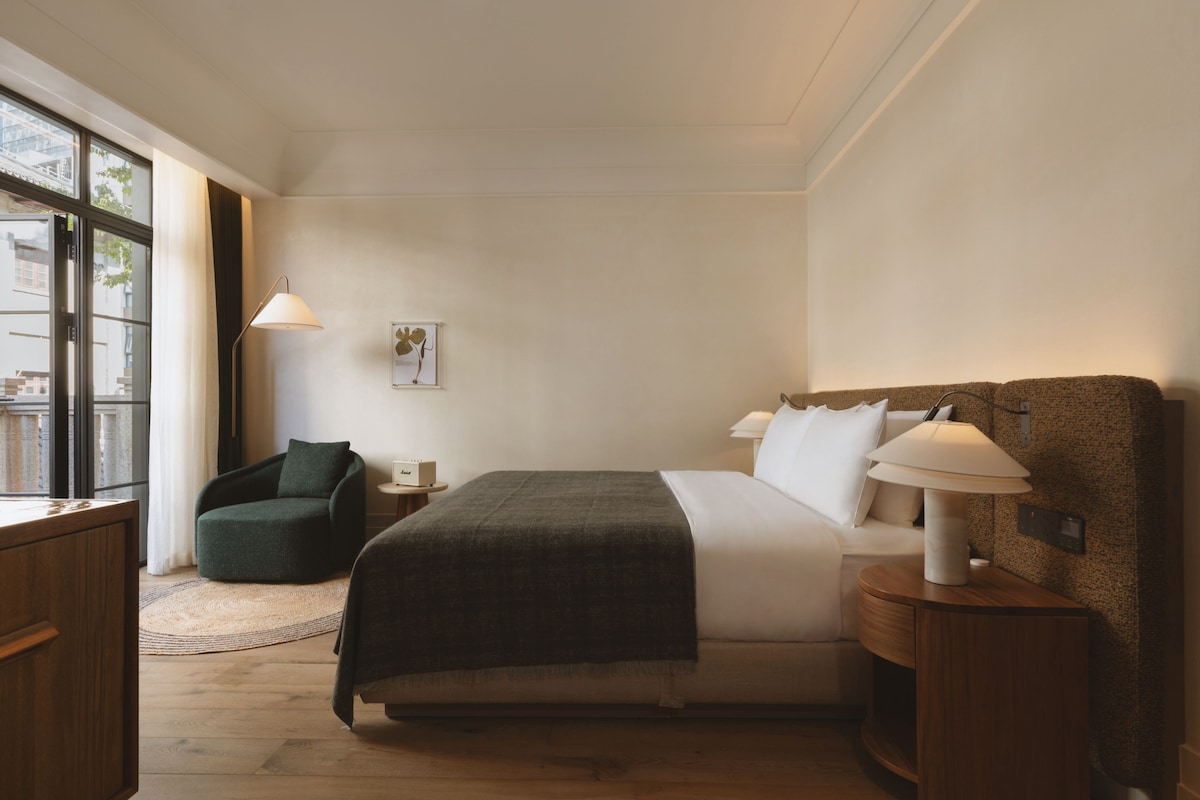 A hotel room with a large bed, bedside lamps, a chair, a small table, and a window with a view of buildings.