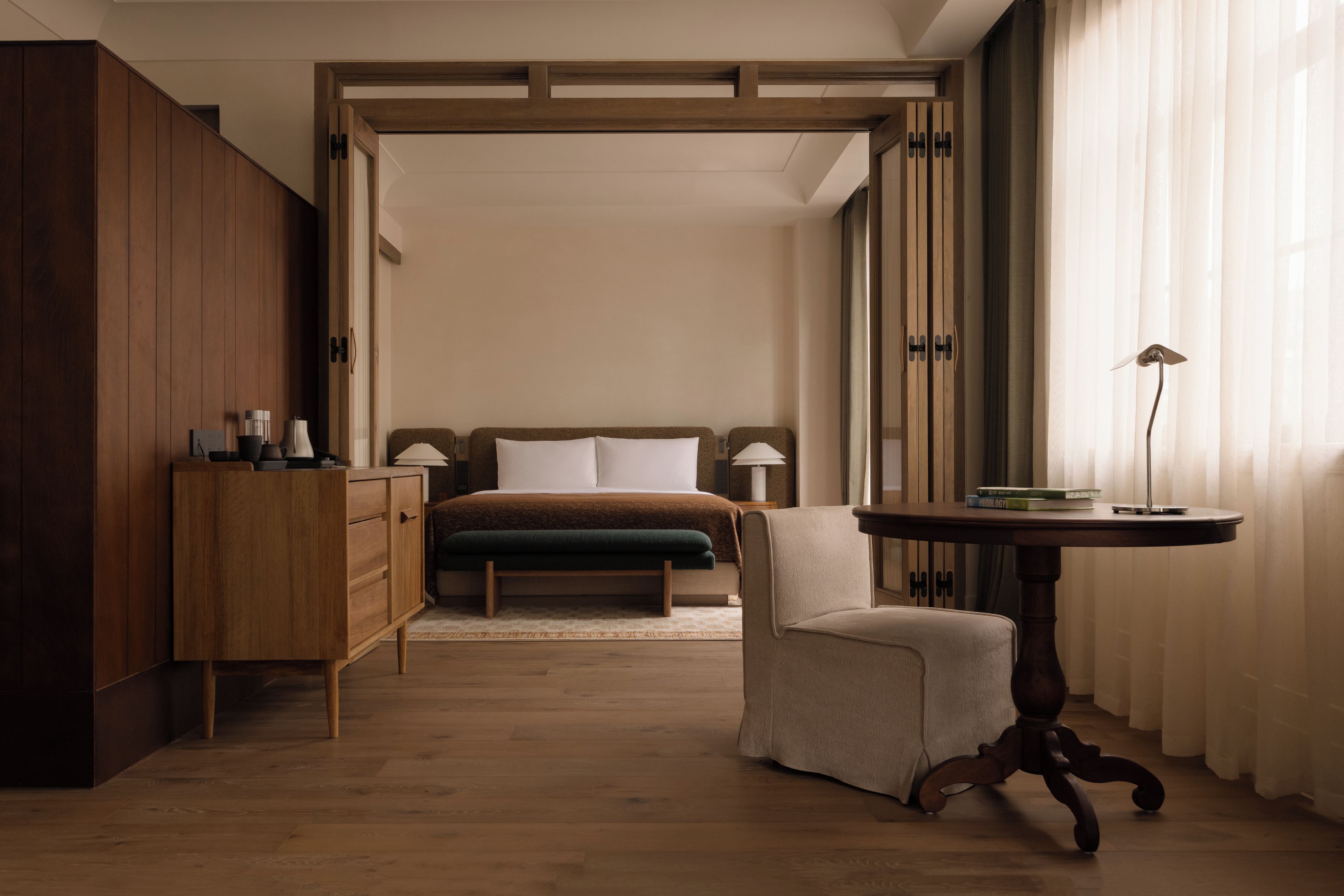 A bedroom with a wooden wardrobe, a bedside table, a chair, a bed with a green blanket, and a bedside lamp.