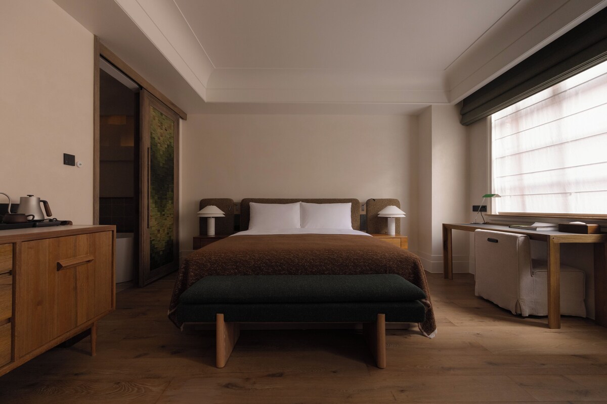 A hotel room with a bed, bedside tables, a desk, and a window.
