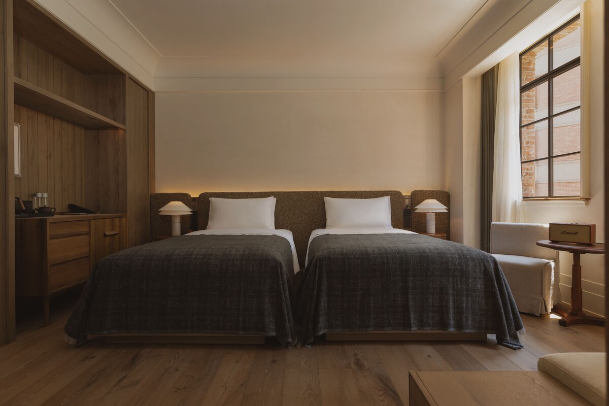 A hotel room with two beds, wooden flooring, a small table with a lamp, and a window with blinds.
