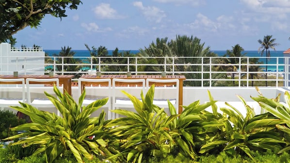 A terrace with white railings, wooden tables, and chairs, overlooking a beach and palm trees.