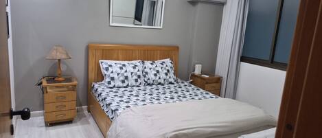 A bedroom with a wooden bed, a bedside table, a lamp, a mirror, and an air conditioning unit.