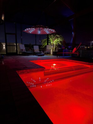 Ein Außenpoolbereich mit einem rot beleuchteten Wasserspiel, Liegestühlen und einem Sonnenschirm.