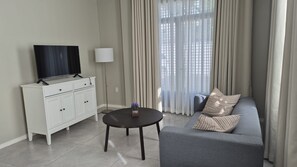 A modern living room with a flat-screen TV on a white cabinet, a round table with a lamp, a grey sofa with striped pillows, and a large window with curtains. - Down town retreat: modern design & premium sleep (Paramaribo)