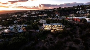 Un edificio residencial moderno de varios pisos con techo plano, rodeado de vegetación densa y otras viviendas.