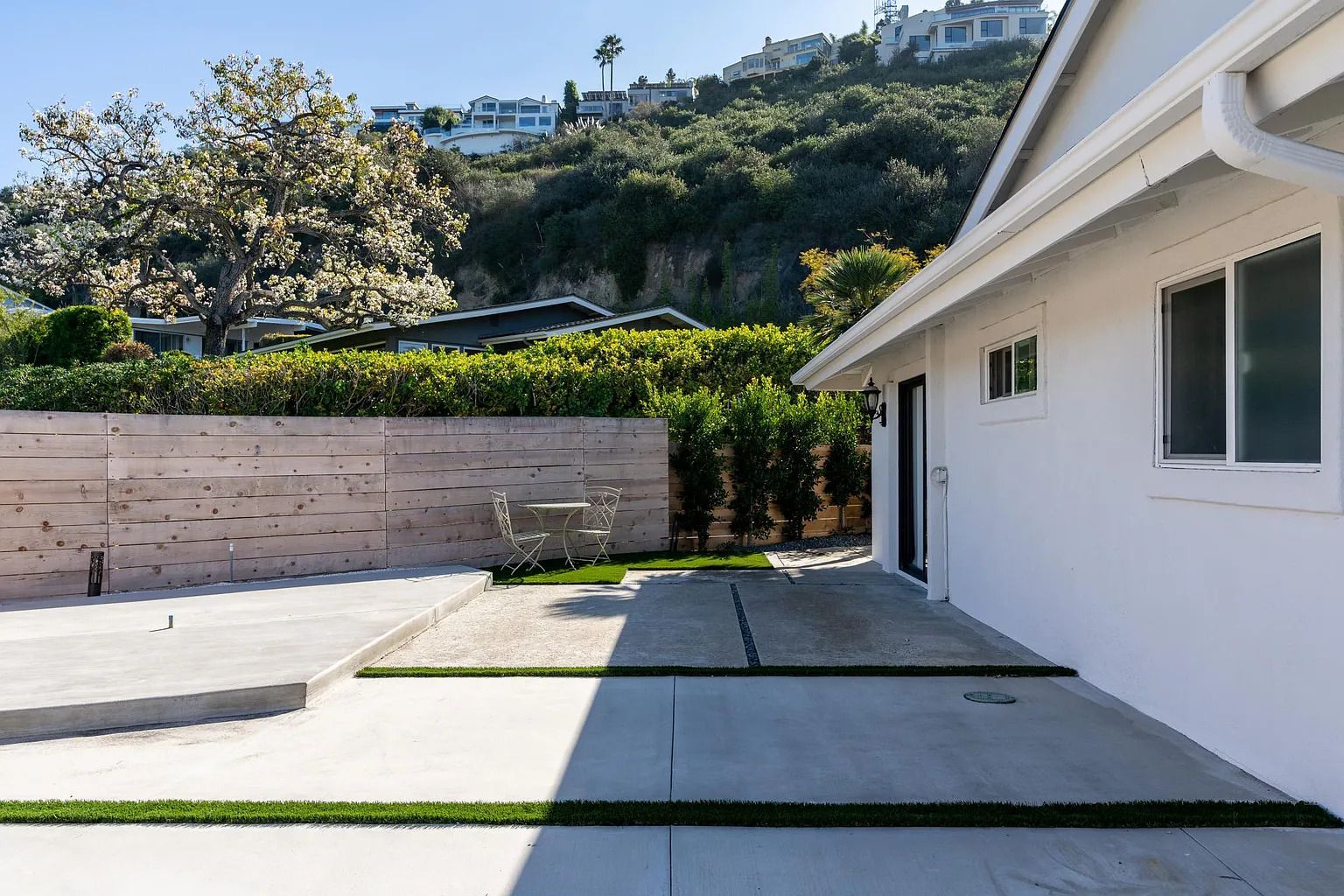 A modern house with a paved patio, a wooden fence, and a view of a hillside with other houses.