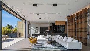A modern living room with a white sofa, a yellow armchair, and a coffee table. There is a large window providing a view of the outdoors. - Bel Air Peristyle By Alora Reserve (Los Angeles)