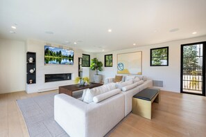 A modern living room with a sectional sofa, a flat-screen TV, a fireplace, and large windows. - Tower Vista By Alora Reserve (Beverly Hills)