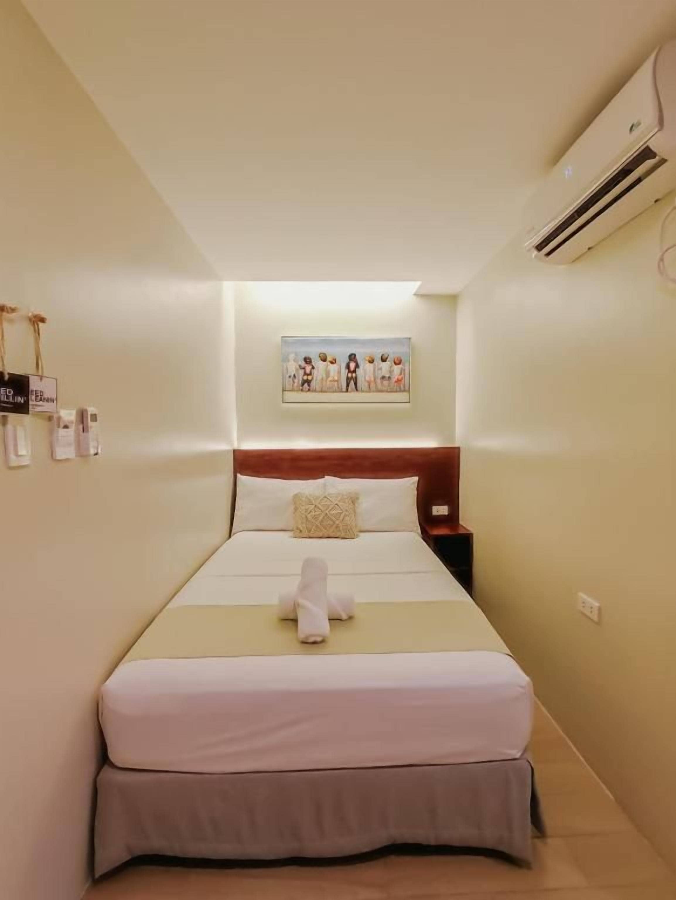 A hotel room with a bed, a nightstand, a wall-mounted air conditioner, and a framed picture above the bed.