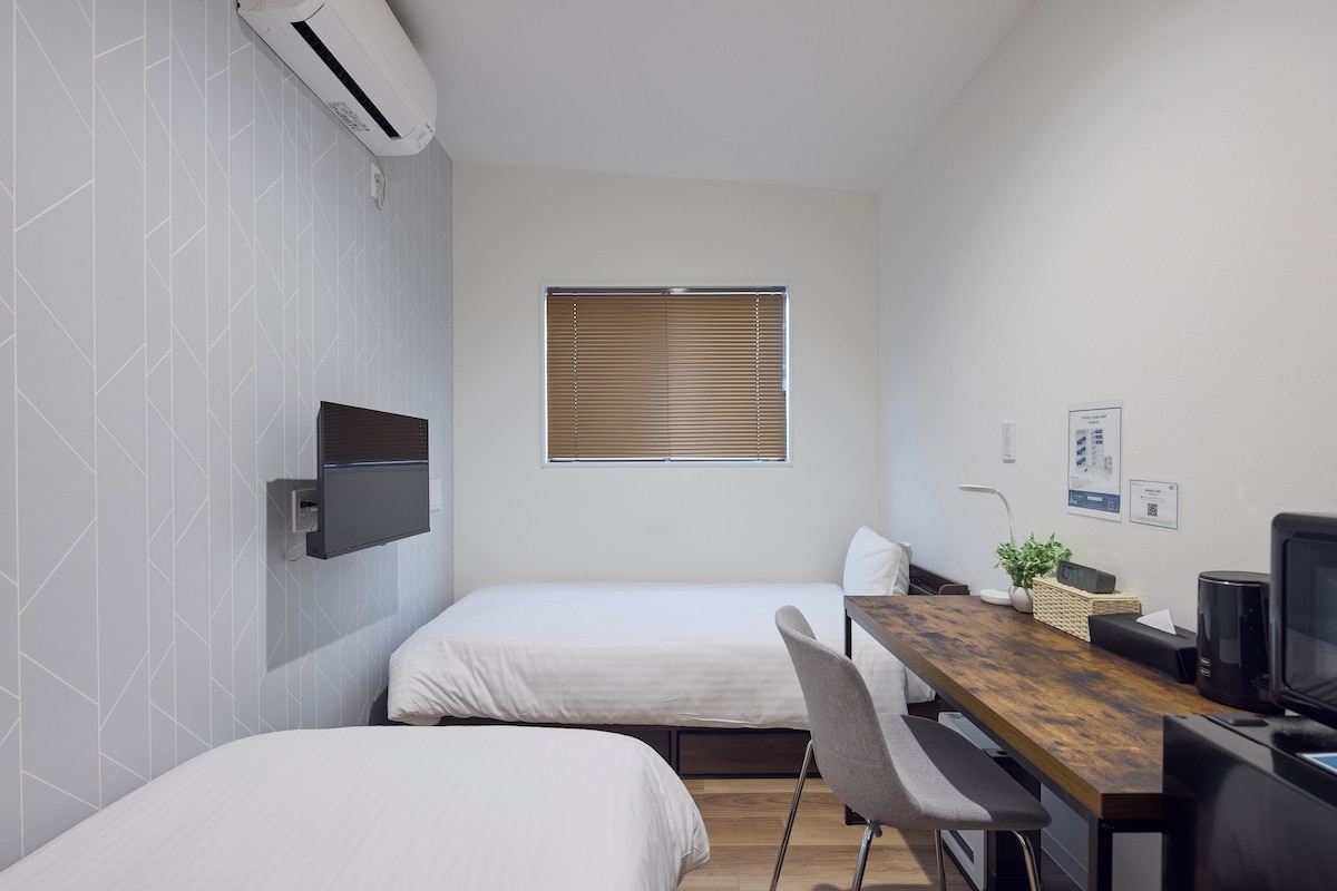 Une petite chambre moderne, équipée de deux lits, d’un bureau en bois, d’une télévision et d’une fenêtre avec des stores.