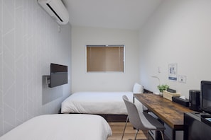 Une petite chambre moderne, équipée de deux lits, d’un bureau en bois, d’une télévision et d’une fenêtre avec des stores.