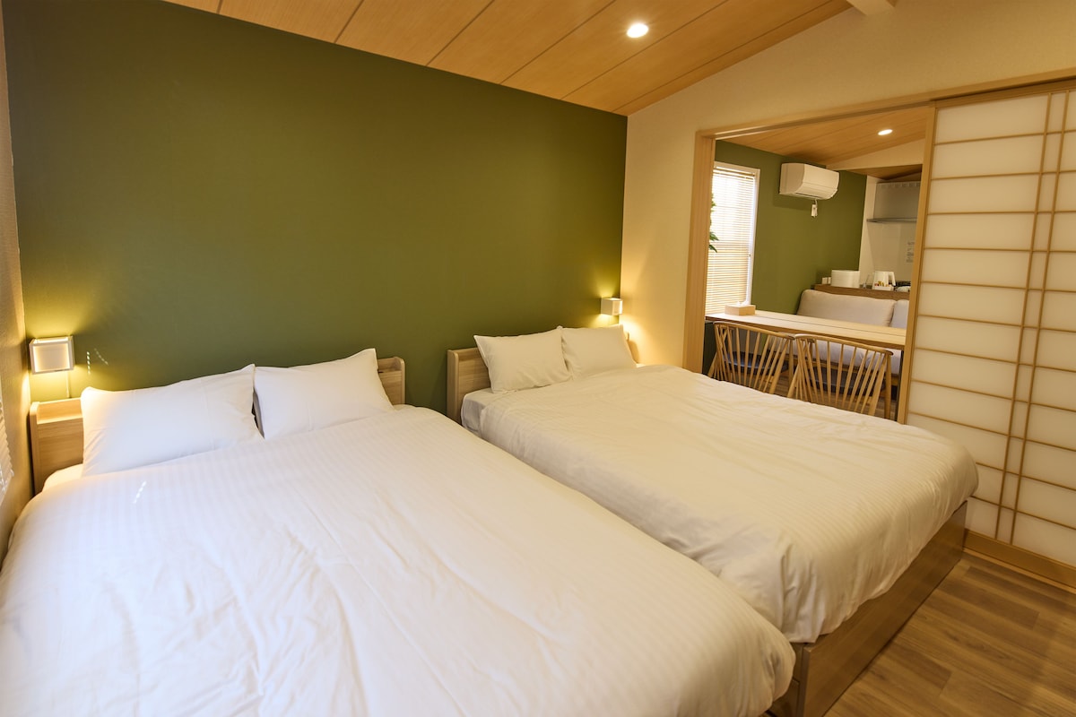 Une chambre d’hôtel avec deux lits, un plafond en bois, un mur vert et un miroir qui reflète la salle de bain.