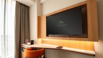 Habitación de hotel con televisor de pantalla plana montado en una pared de madera, un escritorio pequeño con una silla y una ventana con cortinas.