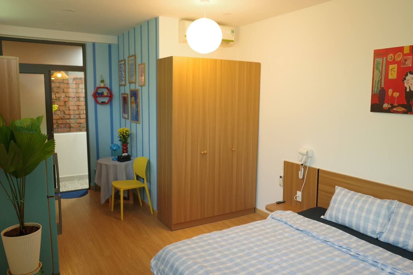 A room with a bed, a wardrobe, a desk with a chair, a potted plant, and a wall with framed pictures.
