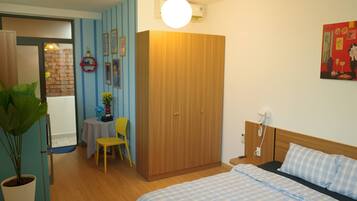 A room with a bed, a wardrobe, a desk with a chair, a potted plant, and a wall with framed pictures.
