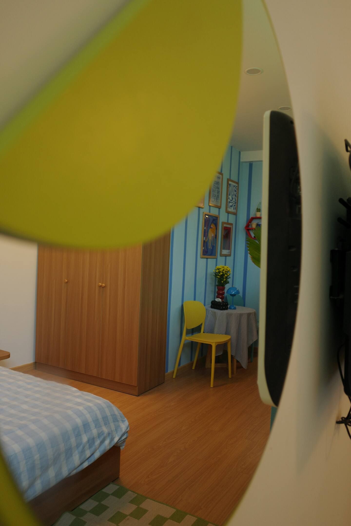 A bedroom with a bed, a wooden wardrobe, a small table with a chair, and a wall with framed pictures.