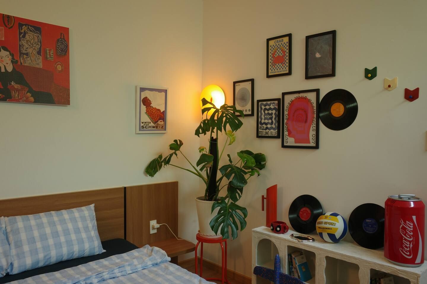 A room with a bed, a potted plant, a shelf with records and a can, and various framed pictures on the wall.