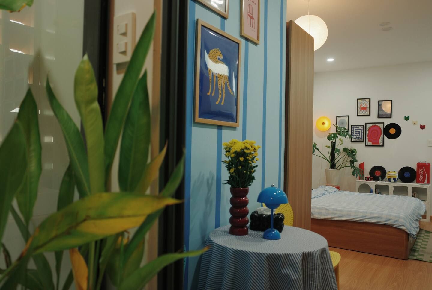 A room with a bed, a round table with a vase of flowers, and a wall with framed pictures and a plant.