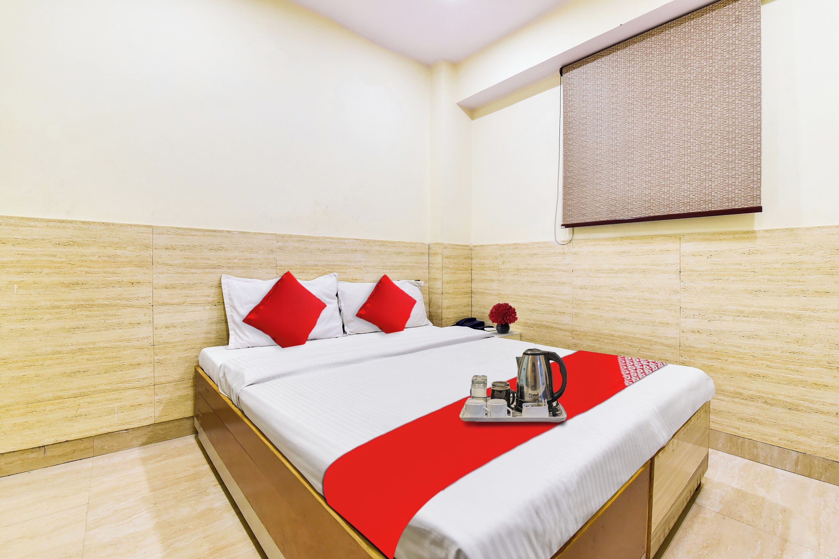 A hotel room with a bed, a red and white bedspread, a red pillow, a vase with flowers, and a coffee maker on the nightstand.