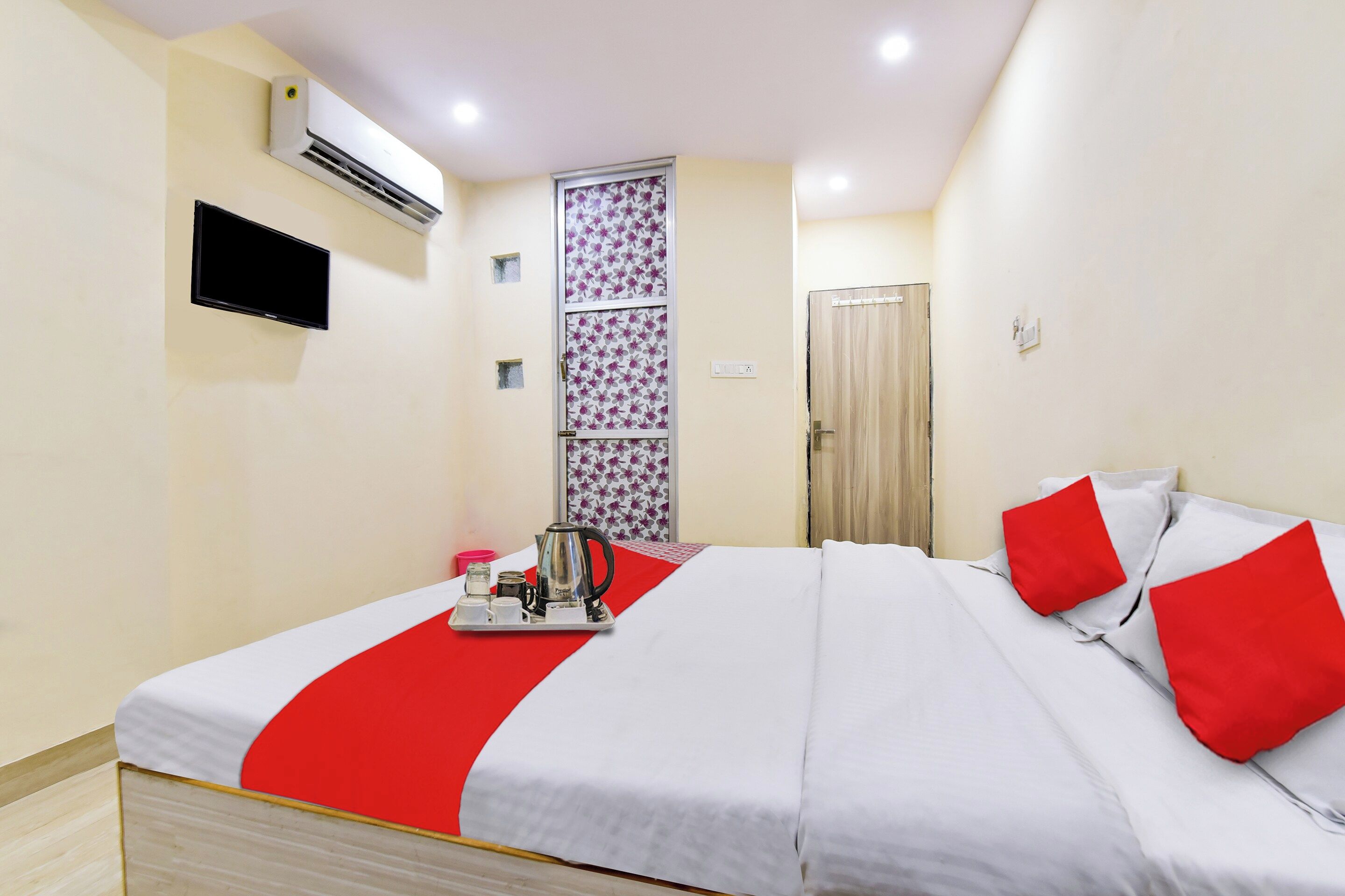 A hotel room with a bed, a TV mounted on the wall, an air conditioning unit, and a door.