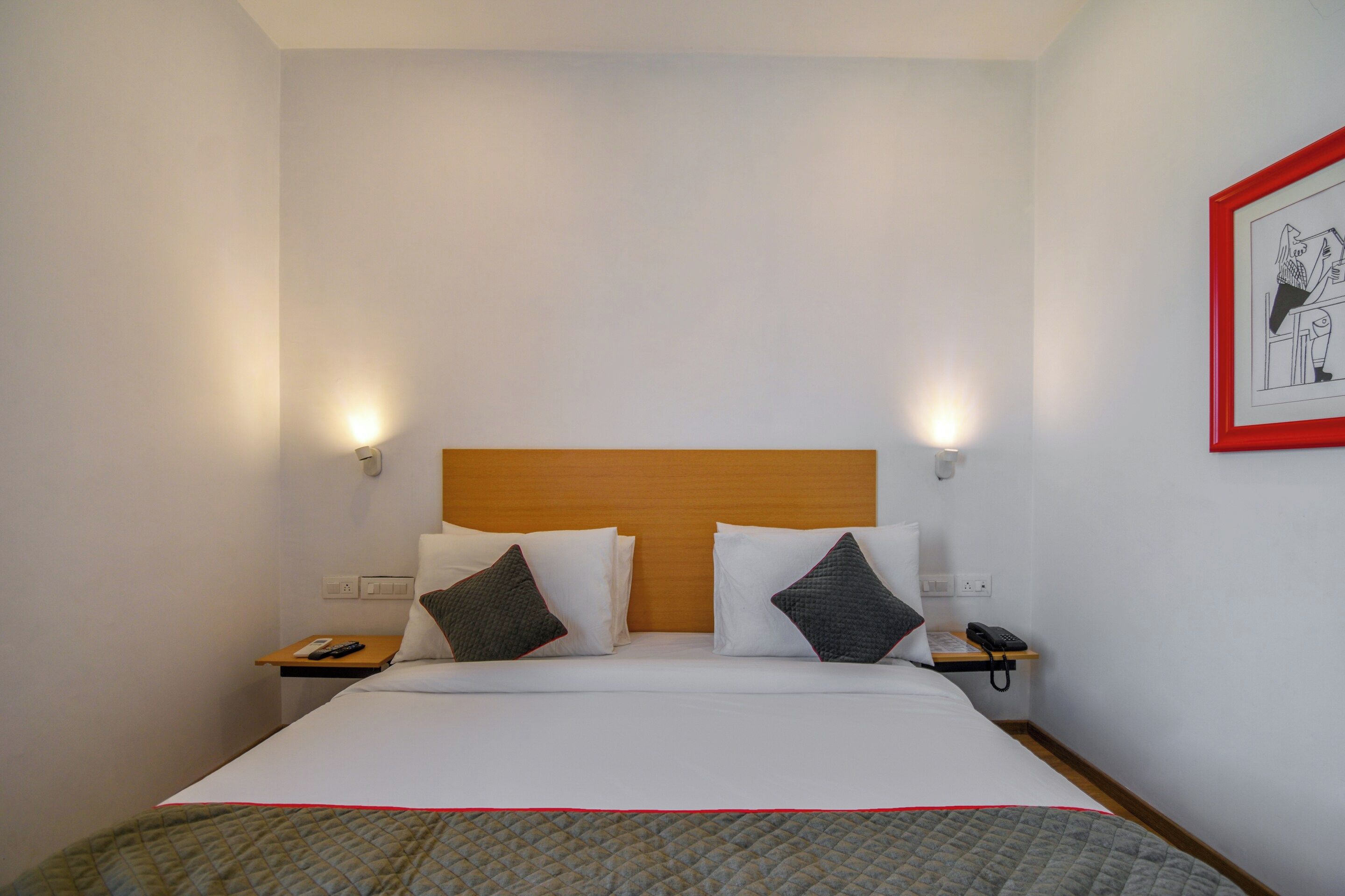 A hotel room with a bed, two bedside tables, a nightstand, a phone, a remote control, and a framed picture on the wall.