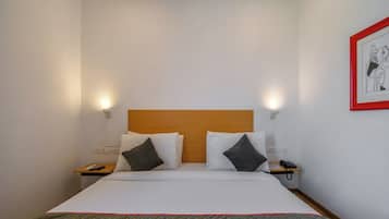 Une chambre d’hôtel avec un lit, deux tables de chevet, une petite table, un téléphone, une télécommande et un tableau encadré au mur.