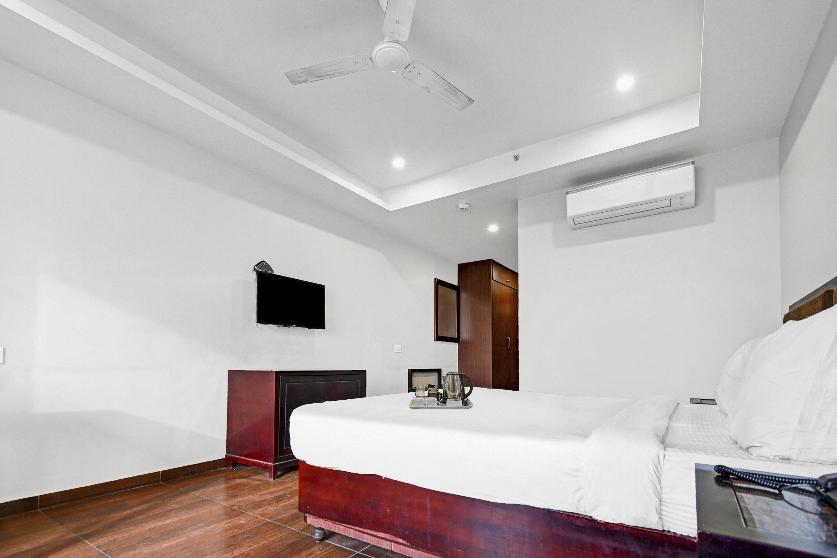 A hotel room with a bed, a flat-screen TV, an air conditioning unit, a wooden cabinet, and a ceiling fan.