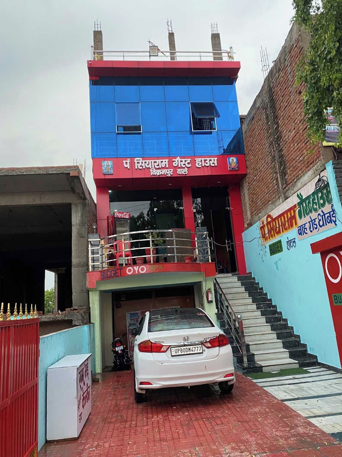 Un bâtiment à la façade bleue et rouge, un escalier menant à une entrée abritée et une voiture blanche garée.