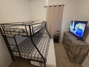 Un lit superposé avec une couvre-lit à motifs, un téléviseur posé sur une commode en bois et un rideau à une fenêtre.