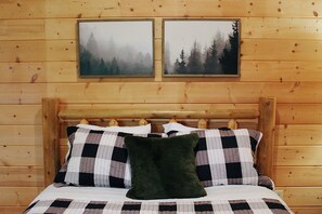 Una cama con colcha a cuadros y almohadas, un cabecero de madera y dos cuadros enmarcados con imágenes de bosques en la pared.