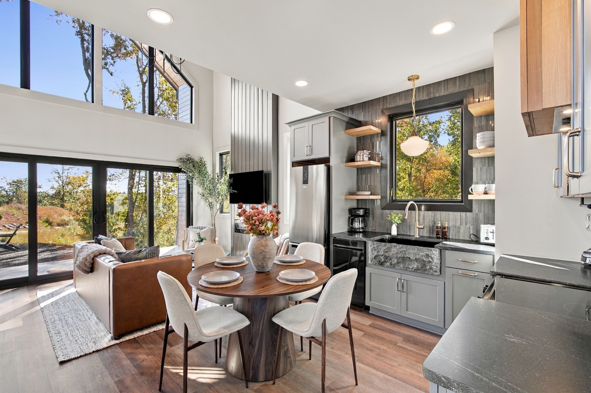 A modern kitchen with a dining area, large windows, and a view of trees.