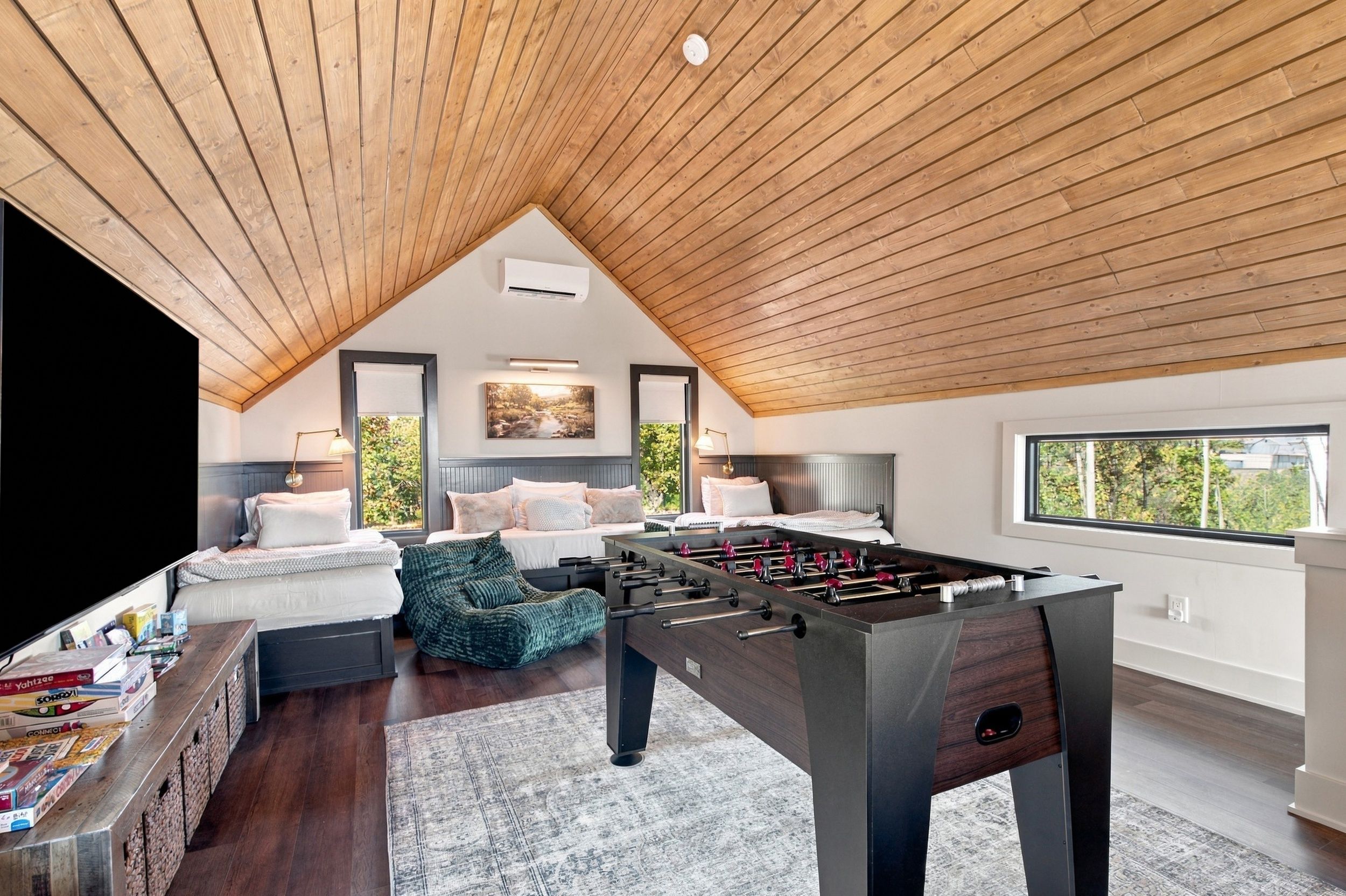A modern loft bedroom with a wooden sloped ceiling, a foosball table, a sofa, and a bed with a view of greenery outside.