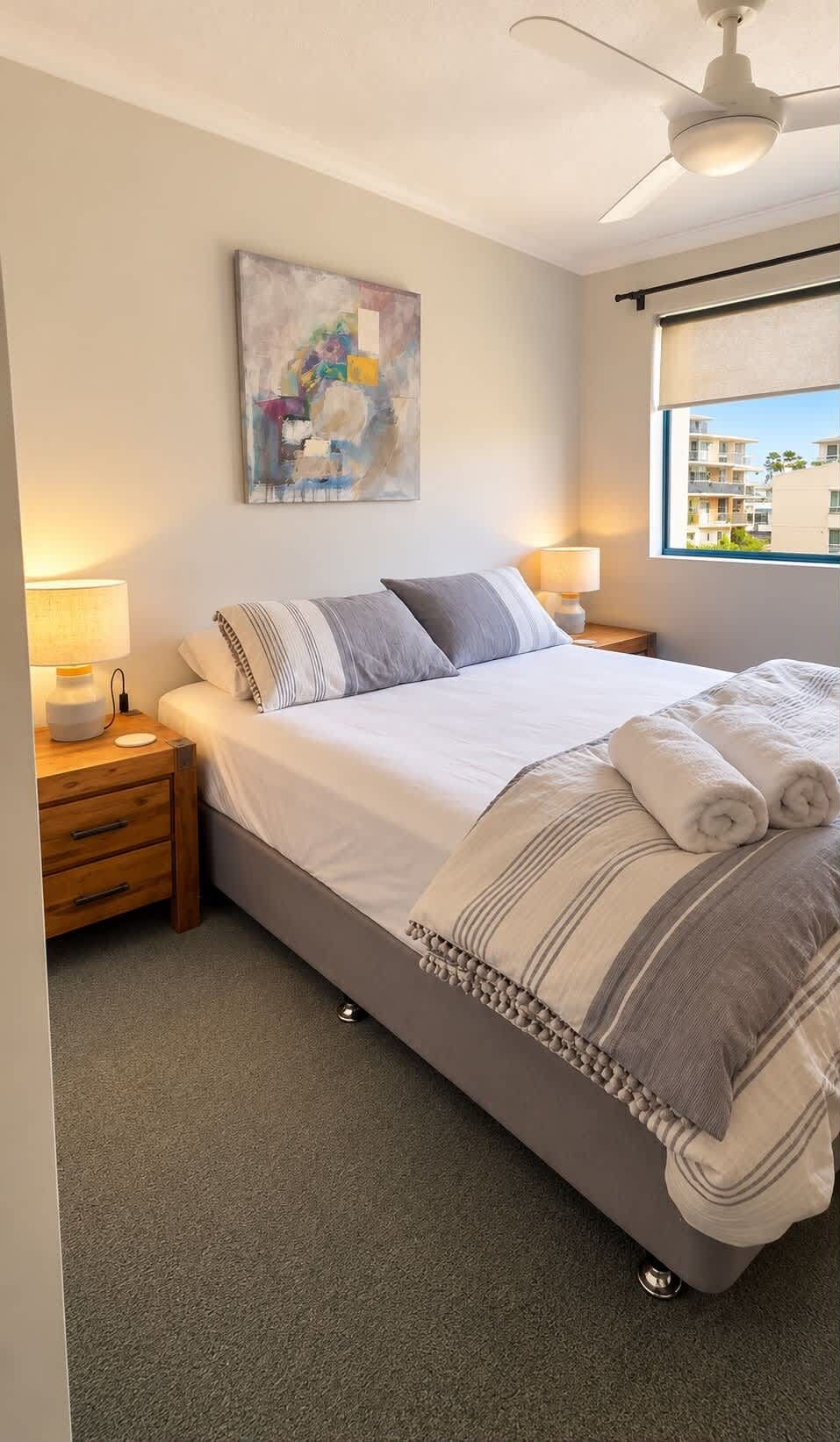 A neatly made bed with pillows and a blanket, a bedside lamp, a wall art piece, and a window with a view of buildings.