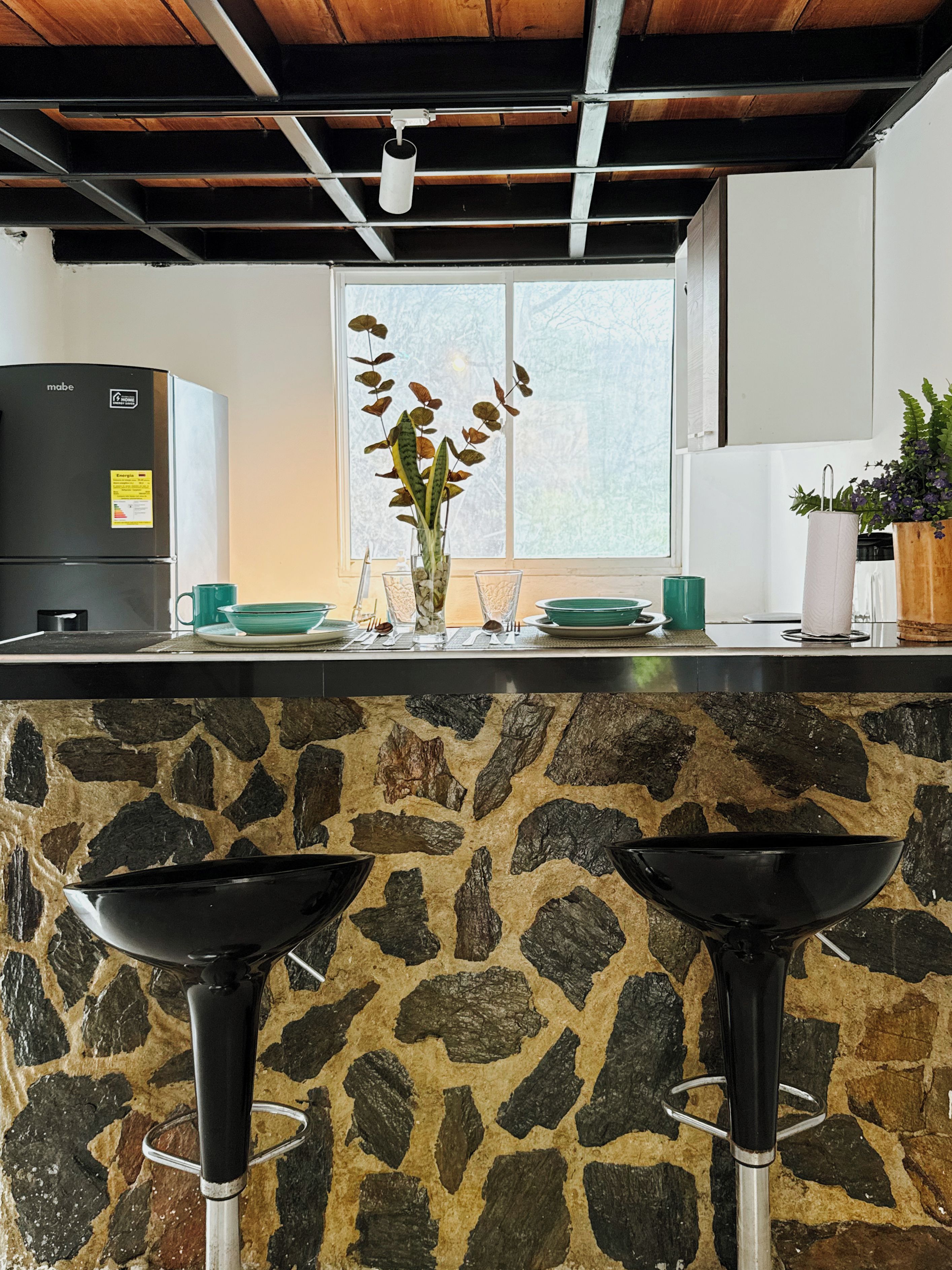 A kitchen with a stone countertop, two black bar stools, a refrigerator, and a window with a view.