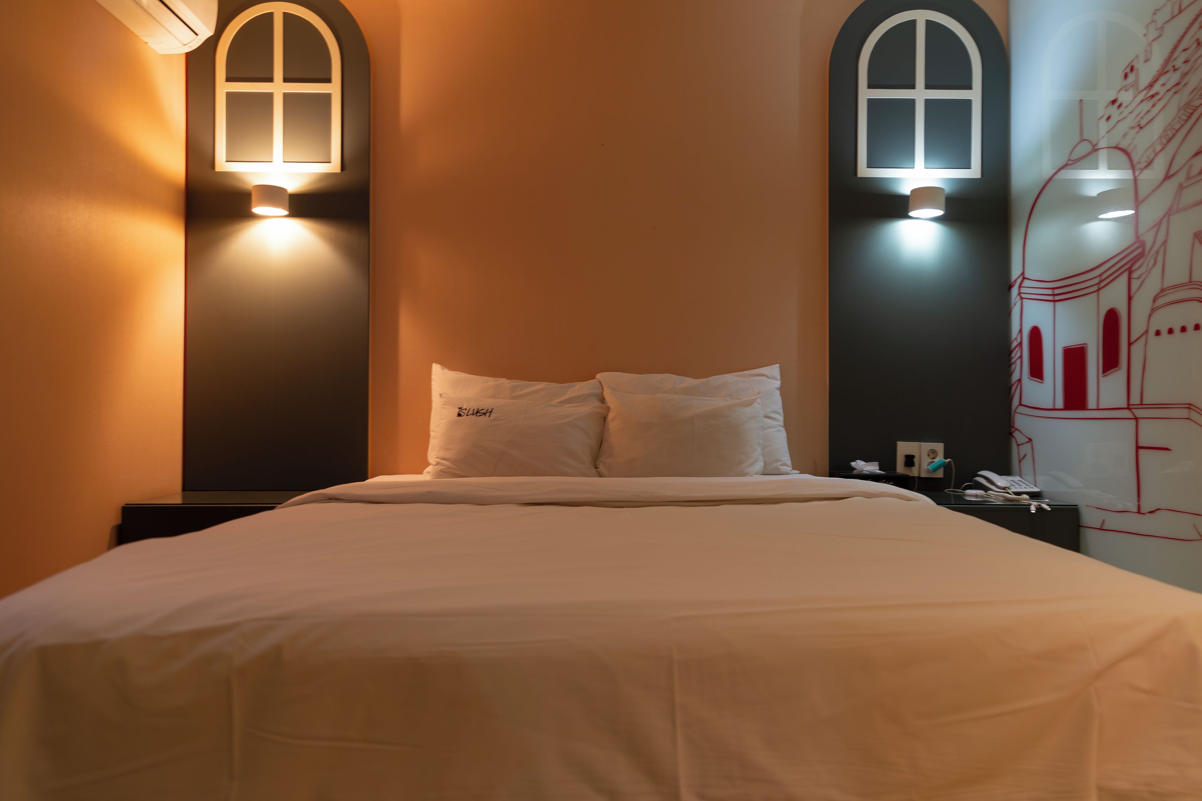 Une chambre d’hôtel avec un lit, deux lampes fixées au mur, une table de chevet et un mur orné d’un dessin linéaire rouge.