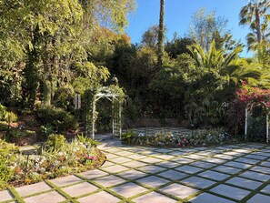 A garden with a paved walkway, a white arbor, and various plants.