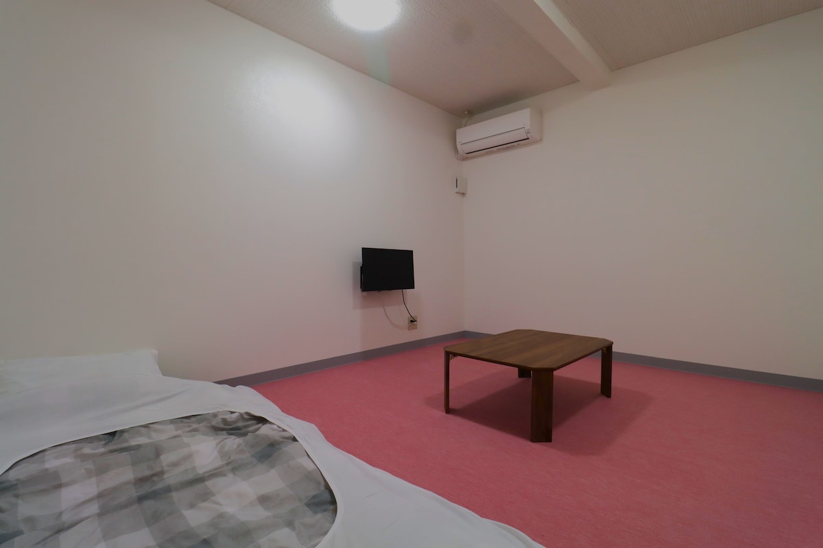 A room with a bed, a small table, an air conditioning unit, and a wall-mounted television.