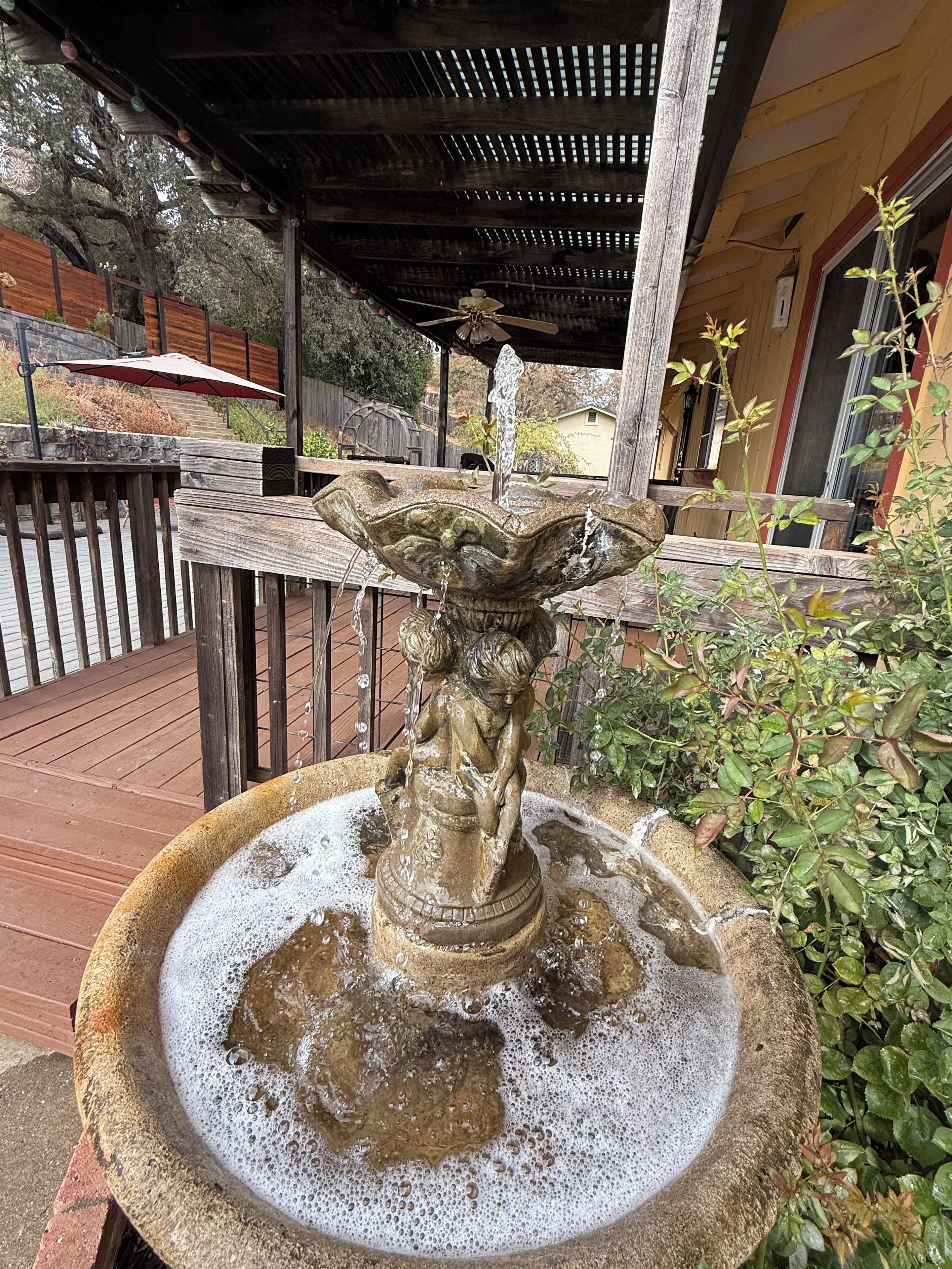 A stone fountain with water spouting from its center, surrounded by a wooden deck and greenery.