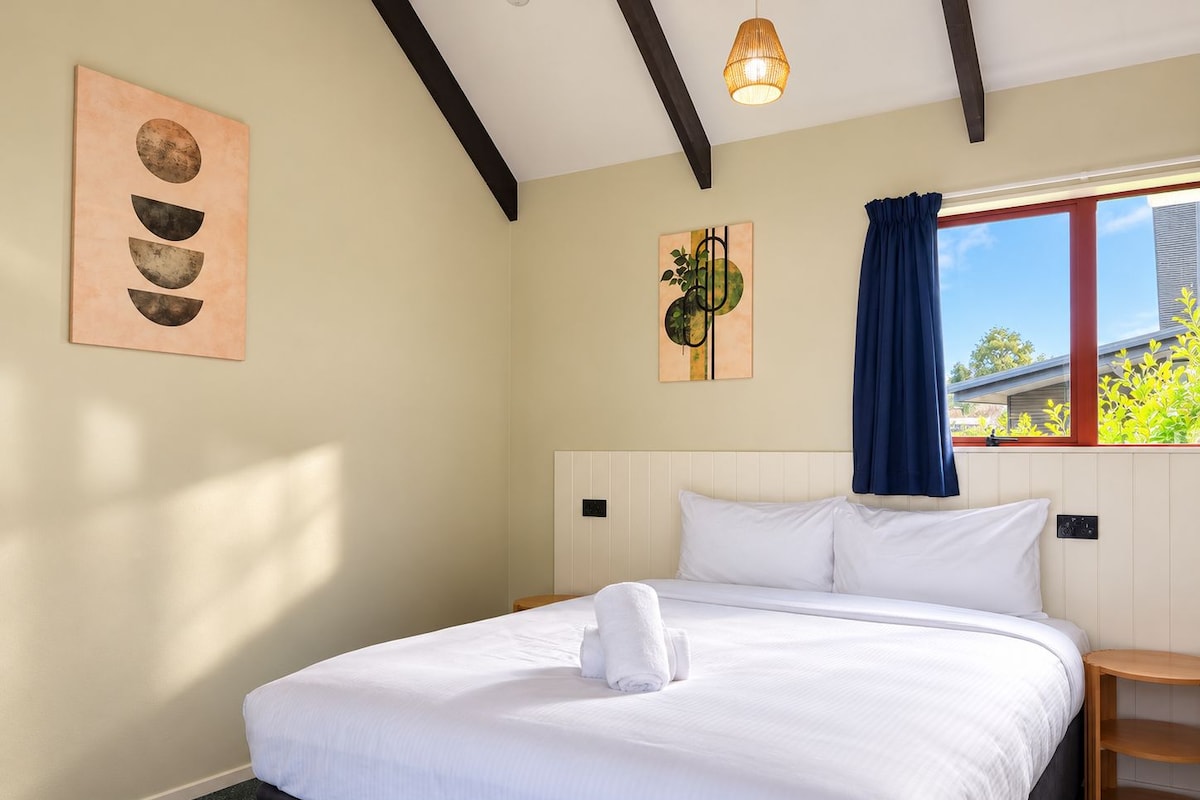 A hotel room with a bed, a window with curtains, a wall-mounted artwork, and a ceiling light fixture.