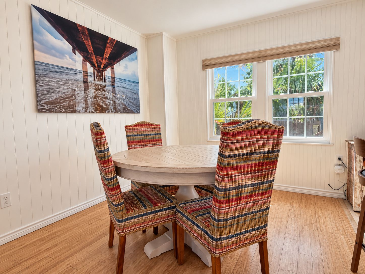 A dining area with a round table and four chairs, a large painting of a pier, and a window with a view of greenery.