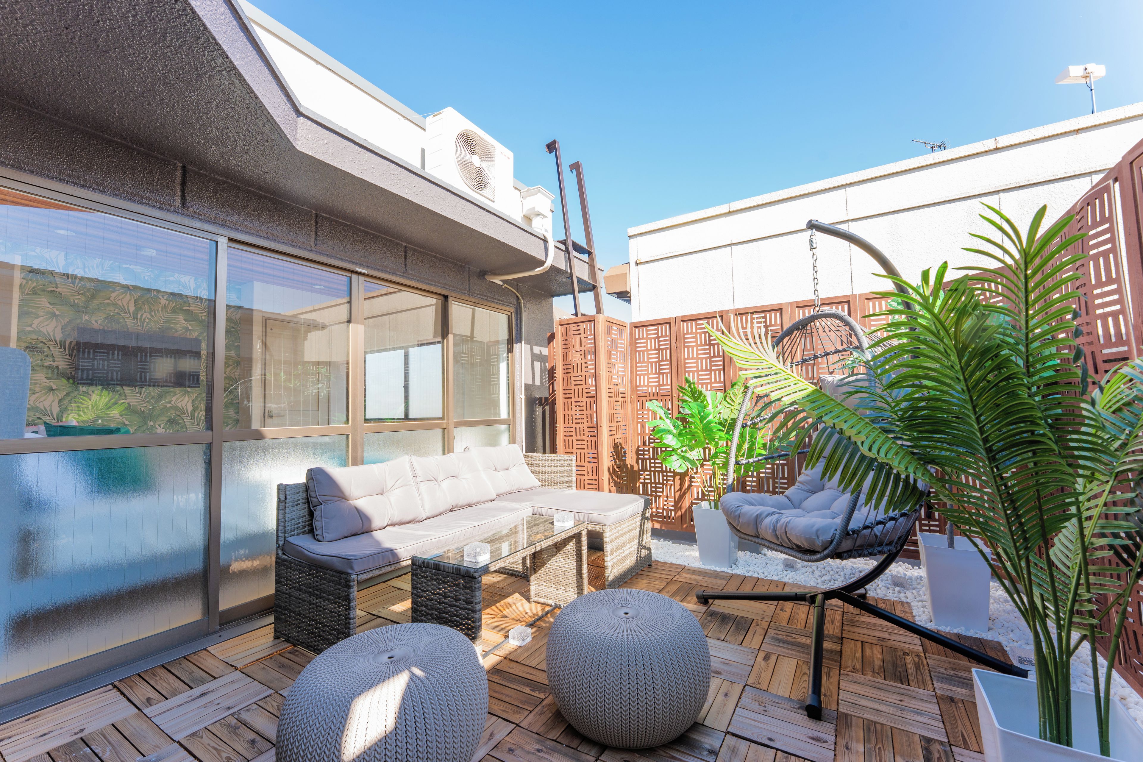 A rooftop patio with a sofa, two round ottomans, a hanging chair, and potted plants.