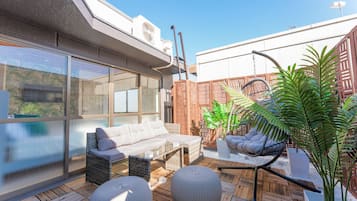 A rooftop patio with a sofa, two round ottomans, a hanging chair, and potted plants.