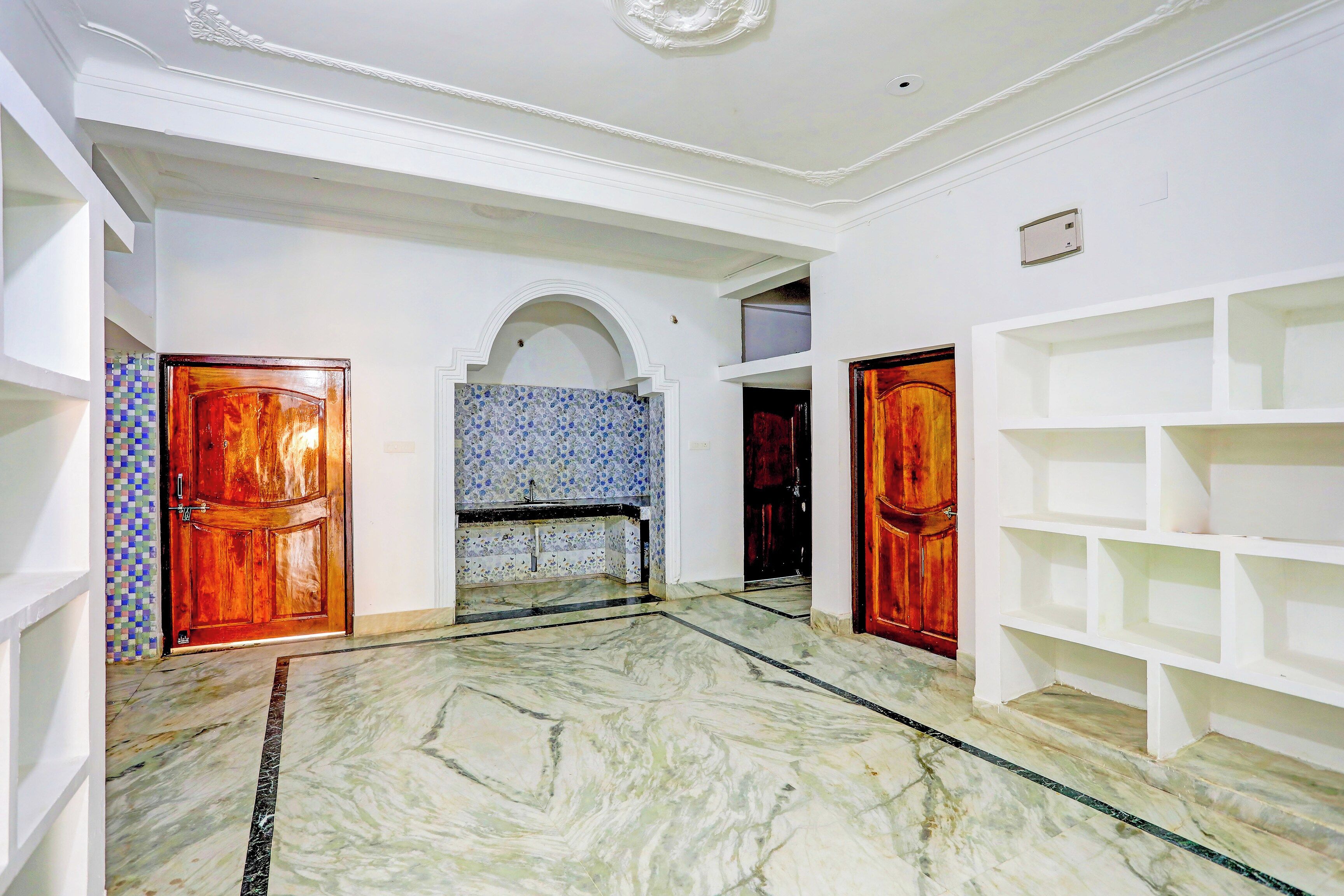 Una habitación amplia con piso de mármol, paredes blancas y salpicadero de azulejos. Se observan dos puertas de madera y un estante blanco.